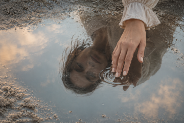 Reflejo difuso de una mujer sobre agua tranquila mientras toca la superficie con la mano en un momento de introspección y sensibilidad interior